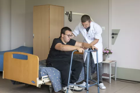2 jeunes en situation d'aide aux personnes, 1 jeune aide son camarade à se lever d'un lit avec un déambulateur 