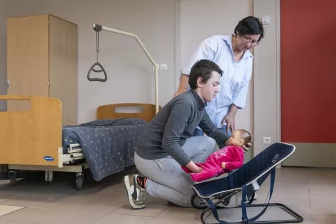 Un jeune et sa professeure en train de poser une poupée dans son fauteuil, cours de puériculture